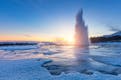 Geysir spewing boiling water into the air during sunset, all surroundings are frozen due to low temperatures in winter.