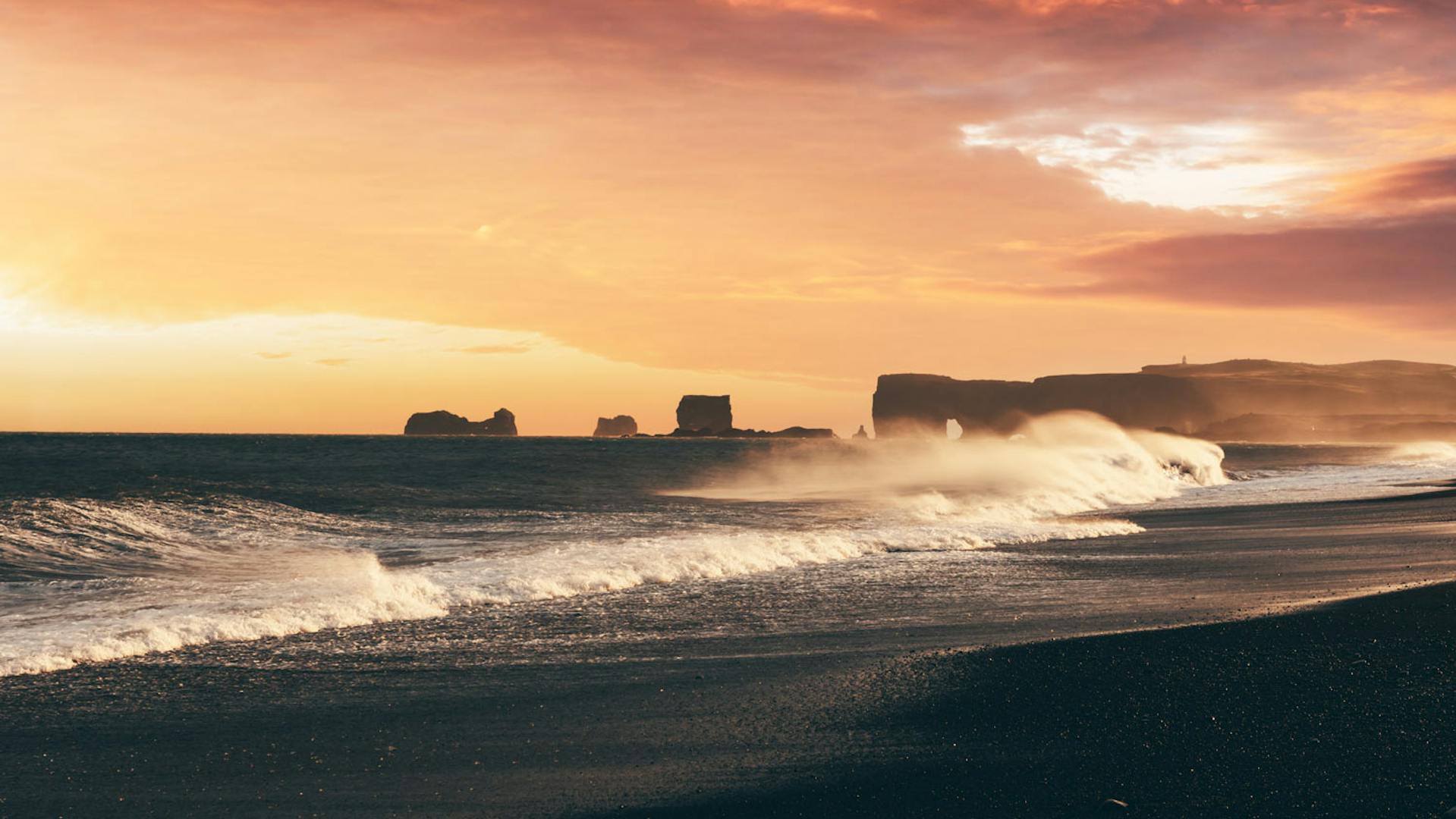 Photo taken from a black sand beach with the wave formation in the foreground, in distance we can observe rock stacks with a lighthouse.