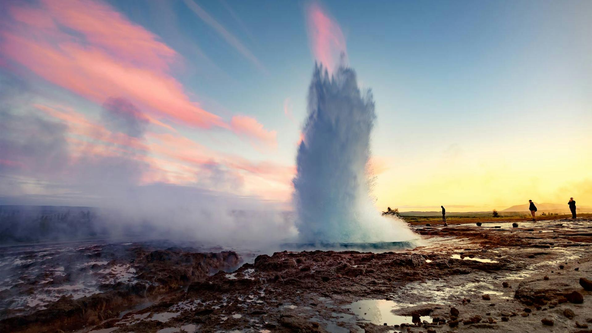Geysir spewing boiling water in the air while people are standing in the surrounding to appreciate this natural phenomenom.
