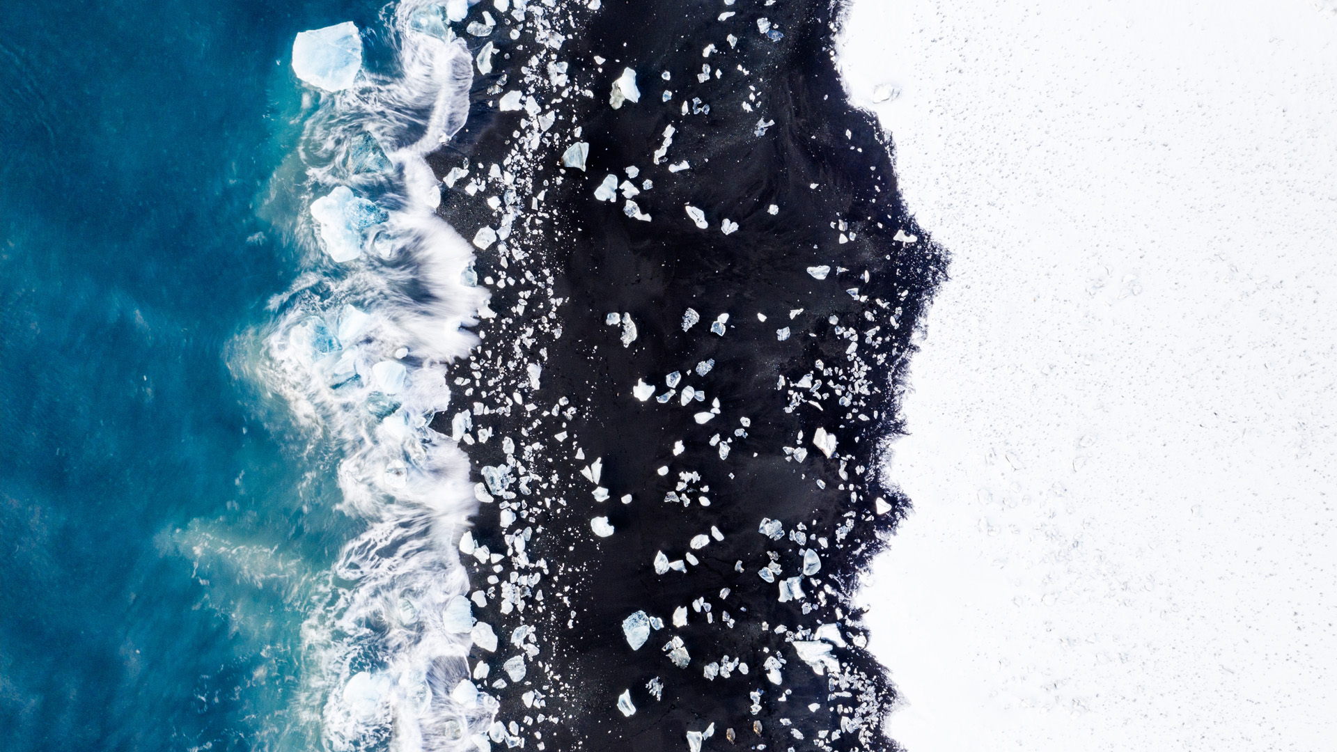 Vertical perspective of a black sand beach during winter time, there is some ice chunks trapped in the snow where the wave breaks and the beach is partially covered with snow.