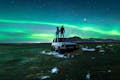 Two people on top of a car in the middle of a grass field with aurora borealis shining in the sky.