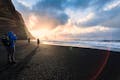 People standing and photographing the sunset on a black sand beach.