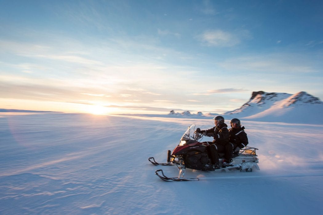 Two people share a snowmobile while they ride over a glacier; the sky is clear blue, the sun is coming up on the horizon, and mountain peaks are in the background.