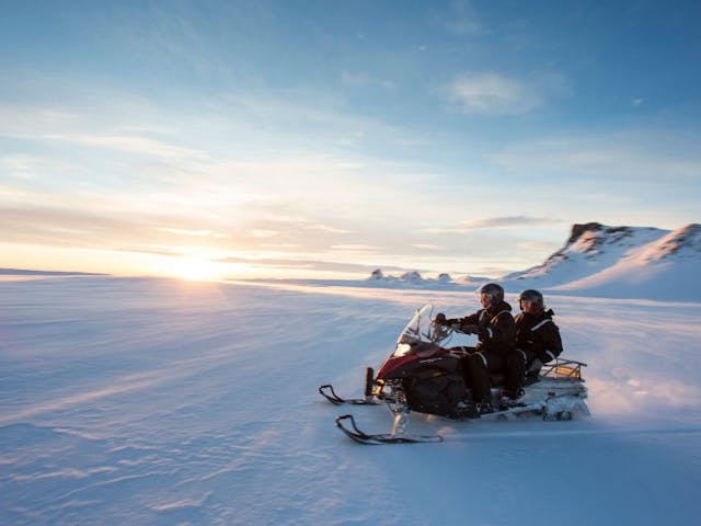 Two people share a snowmobile while they ride over a glacier; the sky is clear blue, the sun is coming up on the horizon, and mountain peaks are in the background.