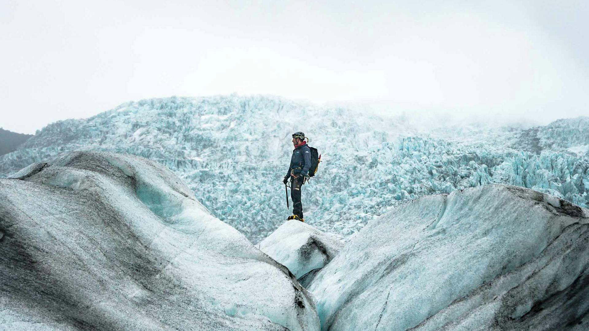 A man standing on top of an ice chunk, behind him it is possible to see glacier formations.