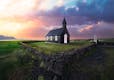 Small stone walls with a black church inside the perimeter, a colorful sunset is striking in background.