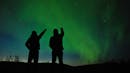 Silhouette of two person point to the sky, while an aurora borealis is showing up.