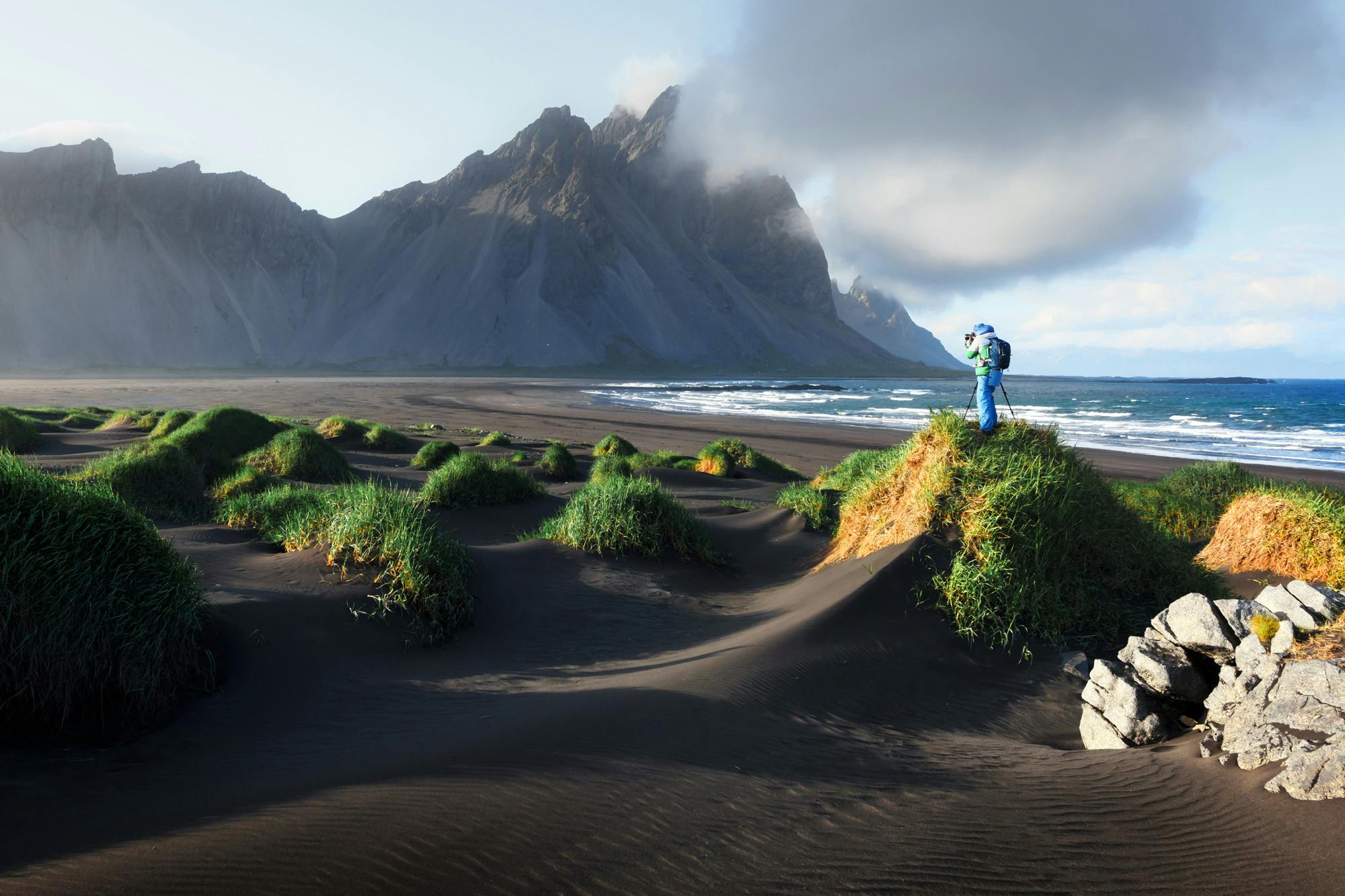 A person photographing the mountain that is located in the distance, the photographer is situated on top of a tuft of herbs, surrounded by black sand.