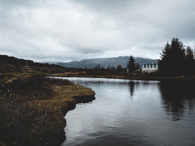 The lake at Þingvellir National Park is still, showing the reflection of the trees, where behind them is a white house with an A-shaped roof that goes in that shape five times. In the background is a mountain and the shore on the other side is mossy green.