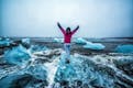 Woman with her arms in the air, standing on top of a little iceberg while waves are crashing on them at a black sand beach, the location is called diamond beach and it's situated in Iceland.
