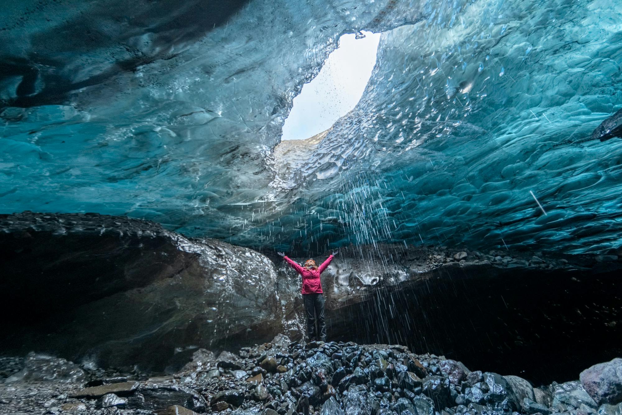 A woman wearing a pink jacket with her arms wide open looking at the ceiling of an ice cave.